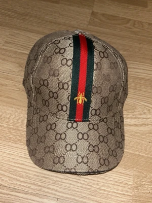 Gucci keps med monogram och ränder - Snygg keps från Gucci i beige med klassiskt GG-monogram över hela ytan. Framtill går en grön och röd rand med ett broderat bi i gult. Justerbar rem baktill. Perfekt accessoar för att lyfta din outfit. OBS FAKE