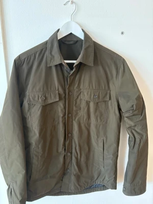 Olivgrön overshirt Tiger of Sweden - Stilren olivgrön overshirt från Tiger of Sweden med två bröstfickor med lock och knappar. Jackan har en klassisk krage, knappstängning framtill och quiltat foder i grått för extra komfort. Perfekt lager på lager-plagg med modern passform.