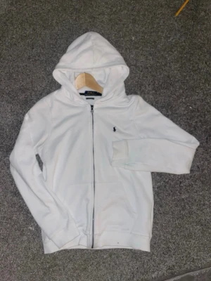 Vit hoodie från Polo Ralph Lauren - Snygg vit hoodie från Polo Ralph Lauren med dragkedja framtill och klassisk huva. Slim fit-modell med diskret broderad logga på bröstet. Perfekt för en clean och stilren look. Mjuk bomullskvalitet och fickor på sidorna.