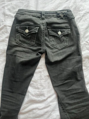 Svarta True Religion jeans - Snygga svarta jeans från True Religion med klassiska kontrastsömmar och stora fickor bak med lock och vita knappar. Jeansen har en rak passform och är tillverkade i ett mjukt denimtyg. Perfekta för dig som gillar streetwear och vill ha en cool look.
