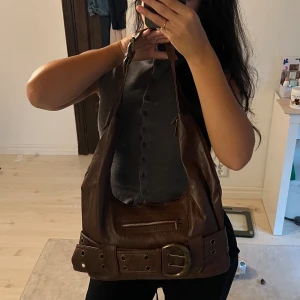 Brun axelremsväska med bältdetalj - Cool brun handväska i mjukt läder med bred axelrem och stor bältesdetalj framtill. Väskan har en dragkedjeficka på framsidan och dekorativa nitar. Perfekt för dig som gillar y2k stil.