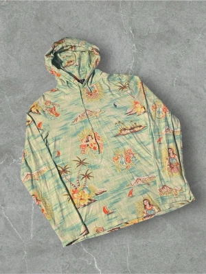 Grön hoodie med Hawaii-mönster Polo Ralph Lauren - Säljer en grön hoodie från Polo Ralph Lauren med färgglatt Hawaii-inspirerat mönster med palmer, blommor och öar. Hoodien har dragkedja framtill och en klassisk huva. Materialet är mjukt och lätt, perfekt för chill dagar.