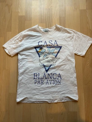 Vit t-shirt från Casablanca med tryck - Vit t-shirt från Casablanca med stort tryck framtill av en svan i blå triangel och texten 'Casa Blanca Par Avion'. Klassisk passform och rund hals. Perfekt för dig som gillar grafiska prints och stilrena färger.