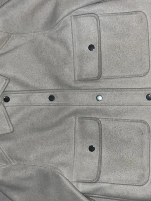 Beige kappa med tryckknappar - Stilren beige kappa med två stora magfickor och silvriga tryckknappar framtill. Jackan har en rak passform och satin på insidan.