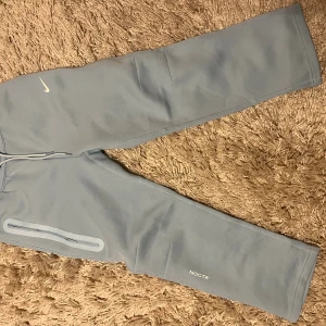Nike NOCTA ljusblå tracksuit - Säljer ett par ljusblå mjukisbyxor från Nike NOCTA-kollektionen. Byxorna har raka ben, dragsko i midjan och en vit Nike-logga på låret. Materialet är mjukt och bekvämt, perfekt för chill dagar. Snygg detalj med NOCTA-tryck på benet och dragkedjeficka på sidan.
