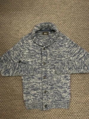 Blåmelerad stickad kofta med knappar - Snygg blåmelerad stickad kofta med stora svarta knappar framtill och hög krage. Koftan har lång ärm och är gjord i ett mjukt garn med blå och vita toner. Perfekt för lager-på-lager och chill dagar.