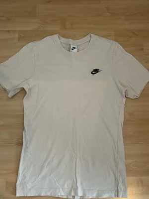 Vit t-shirt från Nike i bomull - Klassisk vit t-shirt från Nike med svart logga broderad på bröstet. Tillverkad i mjuk bomull och har en normal passform med korta ärmar och rund hals. Perfekt basplagg för alla tillfällen.