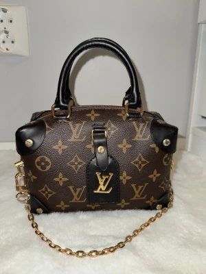  brun monogram handväska - Snygg brun handväska med klassiskt monogrammönster i guld. Väskan har svarta läderdetaljer, guldfärgade metalldelar och en kedjerem. Perfekt storlek för det viktigaste och en ikonisk accessoar för din outfit.