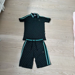 Svarta mönstrade dress från Lacoste - Snygga svart dress från Lacoste med grafiskt rutmönster och gröna samt vita ränder längs sidorna. De har snörning i midjan och en broderad Lacoste-logga framtill. Materialet är mjukt och känns sportigt, perfekt för dig som gillar en chill och trendig look.