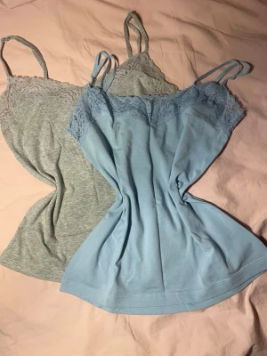 Två spetslinnetoppar från Esmara - Säljer två snygga linnetoppar från Esmara i ljusblått och ljusgrått. Båda har tunna axelband och spetsdetaljer upptill. Materialet är mjukt och stretchigt med bomull och elastan, perfekt för lager på lager eller sommaroutfits.