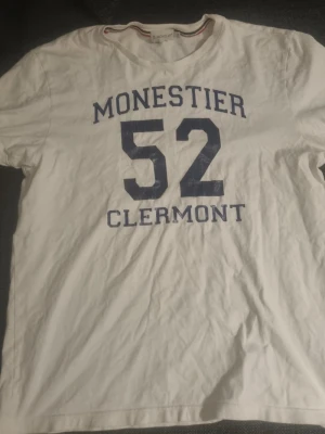 Vit Moncler t-shirt med tryck - Vit t-shirt från Moncler med blått tryck 'Monestier 52 Clermont' på framsidan. Klassisk passform och rund halsringning. Moncler-logga på ärmen. Perfekt för en avslappnad och sportig stil.