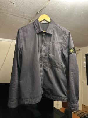 Mörkblå overshirt från Stone Island - Snygg mörkblå overshirt från Stone Island med klassisk patch på ärmen. Jackan har dragkedja framtill, bröstficka med knapp som passar till alla typer av outfits. Den är autentisk och kvitto finns då den är köpt på NK i Stockholm för 4800 kr. Använd väldigt sparsamt. Vid frågor eller funderingar så är det bara att höra av dig! 