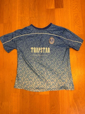 Blå sporttröja från Trapstar - Snygg blå sporttröja från Trapstar med vit logga och text framtill samt stort nummer 22 på ryggen. Tröjan har ett mönster i olika blå nyanser, rund halsringning och vita detaljer längs axlarna. Perfekt för dig som vill sticka ut på träningen.