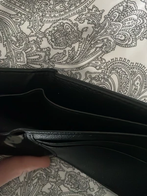 Svart klassisk plånbok i läder - Snygg svart plånbok i slimmad design med flera kortfack och ett genomskinligt fönster för ID eller körkort. Tillverkad i slätt läder och har plats för både sedlar och kort. Perfekt för dig som vill ha en stilren och praktisk accessoar.