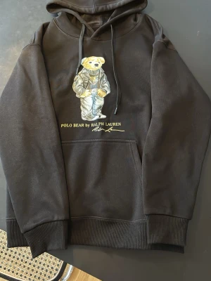 Svart hoodie Polo Bear Ralph Lauren - (Pris kan diskuteras)! Svart hoodie från Ralph Lauren med den ikoniska Polo Bear-björnen broderad på bröstet. Hoodien har huva med dragsko och stor magficka framtill. Tillverkad i mjukt bomullsmaterial och har ribbade muddar vid ärmar och nederkant. Perfekt för en avslappnad och trendig look.