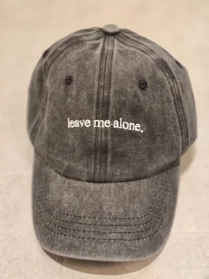 Svart keps med text 'leave me alone' - Cool svart keps i tvättad bomull med vit broderad text 'leave me alone.' framtill. Justerbar rem med metallspänne baktill för perfekt passform. Perfekt accessoar för en avslappnad streetstyle-look.