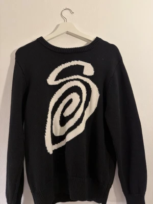 Svart stickad tröja från Stussy - Svart stickad tröja från Stussy med ett stort vitt spiraltryck på framsidan. Tröjan har rund halsringning och ribbade muddar vid ärmslut och nederkant. Perfekt för dig som gillar streetwear och vill sticka ut med en cool design.