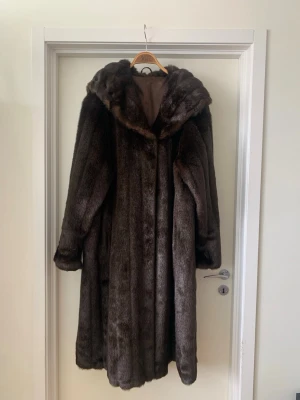 Lång mörkbrun pälsjacka med krage - Säljer en lång och lyxig mörkbrun vintage pälsjacka med bred krage och hel längd. Jackan har en klassisk rak passform och är fodrad på insidan. Perfekt för kalla vinterdagar när du vill hålla dig varm och snygg. Mått i cm: 128 längd, axelvidd 85, ärm 42. Modellen är ca 170cm lång