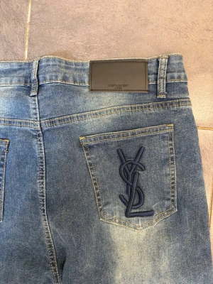 Blå jeans från Saint Laurent - Snygga blå jeans från Saint Laurent med klassisk femficksmodell och broderad YSL-logga på bakfickan. Jeansen har en tvättad look med ljusare partier framtill och bälteshällor. Perfekta för en trendig och avslappnad stil.