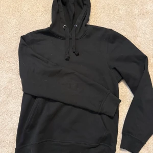 Svart hoodie med huva och ficka - En klassisk svart hoodie med huva och dragsnören. Tröjan har en stor magficka framtill och ribbade muddar vid ärmslut och nederkant. Perfekt för en avslappnad och cool look. Materialet känns mjukt och bekvämt.