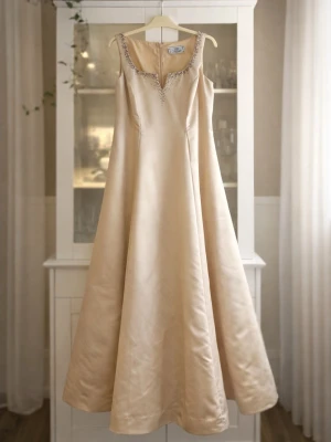 Exklusiv balklänning i champagne – John Charles Collection! - En otroligt elegant och sofistikerad golvlång balklänning i vacker champagnefärgad satin från John Charles Collection.  Klänningen har en smickrande A-linjeform som markerar midjan och faller ut i en lyxig, följsam kjol. Den djupa men stilrena urringningen är dekorerad med handsydda pärlor och stenar som ger ett exklusivt och tidlöst intryck.  Perfekt för dig som vill bära något klassiskt, feminint och riktigt elegant på bal, bröllop eller annan högtidlig tillställning.  Märke: John Charles 