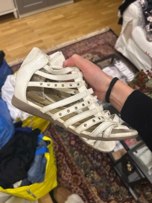 Vita gladiatorsandaler från Din Sko - Snygga vita gladiatorsandaler från Din Sko med flera smala remmar och små guldfärgade nitar. Sandalerna har öppen tå och häl, samt dragkedja bak för enkel påtagning. 