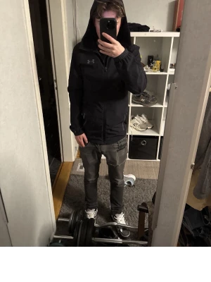 Svart vindjacka från Under Armour - Säljer en svart vindjacka från Under Armour med huva och dragkedja framtill. Jackan har diskret logga på bröstet och praktiska fickor med dragkedja. Perfekt för att hålla vinden borta och ge en sportig look.