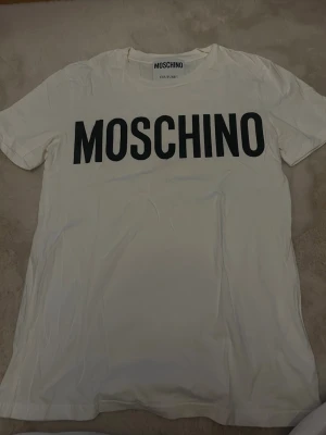 Vit Moschino t-shirt med tryck - Säljer en vit t-shirt från Moschino med stort svart logotryck över bröstet. Köpt på nk på herravdelningen, använd ett fåtal gånger!