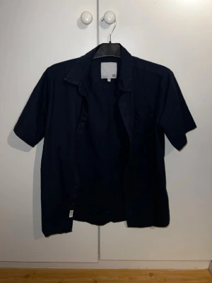 Mörkblå kortärmad skjorta från H&M - Säljer en stilren mörkblå skjorta från lager157 med korta ärmar och klassisk krage. Skjortan är tillverkad i ett lätt bomullsmaterial och har en enkel design utan mönster. Perfekt för dig som gillar en clean och enkel look.