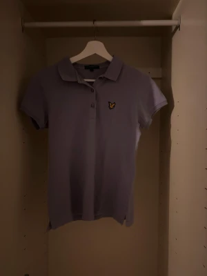 Ljuslila pikétröja från Lyle & Scott - Snygg ljuslila pikétröja från Lyle & Scott med klassisk krage, korta ärmar och knappslå framtill. Tröjan har den ikoniska gula örnloggan broderad på bröstet. Perfekt till jeans eller kjol för en avslappnad men stilren look. M men sitter som S