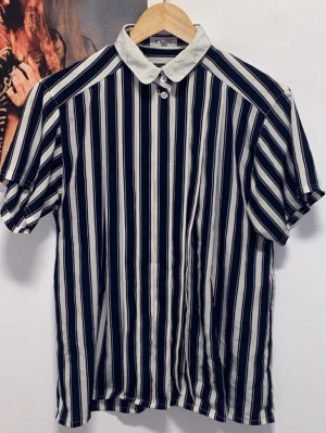 Vintage JOBIS Clothing skjorta - Snygg vintage skjorta från JOBIS Clothing med vertikala svarta och vita ränder och kontrasterande vit krage. Skjortan har korta ärmar och knäppning framtill. Materialet känns lätt och luftigt, super kvalité. Lite oversize så skulle även kunna passa storlek 38-40.