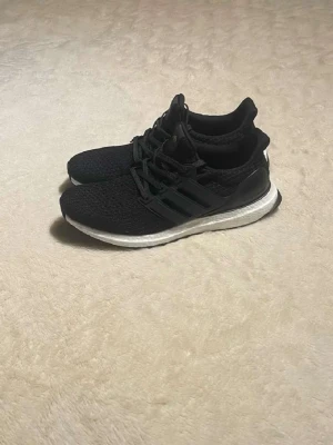 Svarta Adidas Ultra Boost sneakers - Säljer ett par svarta Adidas Ultra Boost sneakers med stickad ovandel och vita Boost-sulor. Skorna har klassiska tre ränder på sidan, svart snörning och en sportig, bekväm design. Perfekta för dig som gillar streetwear och vill ha riktigt sköna sneakers.