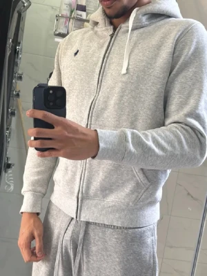 Ljusgrå zip hoodie från Polo Ralph Lauren - Snygg ljusgrå hoodie med dragkedja från Polo Ralph Lauren. Klassisk design med huva och vita dragsnören, två fickor framtill och broderad logga på bröstet. Tillverkad i mjuk bomullsblandning som är perfekt för chill dagar. Passar S och M. Ny