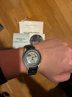 Omega x Swatch Speedmaster klocka - Säljer en Omega x Swatch Speedmaster MoonSwatch med svart armband och silverfärgad urtavla. Klockan har flera subdials och tydliga markeringar, samt texten 'Speedmaster' på armbandet. En riktigt snygg och ikonisk klocka för dig som gillar stilrena accessoarer.1:1