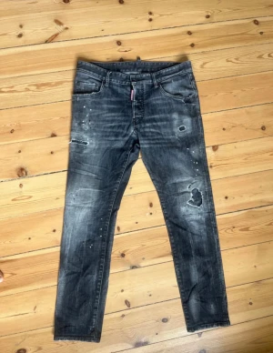 Äkta Dsquared2 jeans - Säljer ett par svarta Dsquared2 jeans med coola slitningar och färgstänk för en edgy look. Jeansen har raka ben, klassisk femficksmodell och tydliga detaljer som trasiga partier och tvättad finish. Perfekta för dig som gillar streetwear och vill sticka ut.