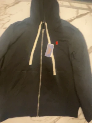 Svart hoodie från Polo Ralph Lauren - Svart hoodie med dragkedja från Polo Ralph Lauren. Klassisk design med vit dragsko i huvan och röd broderad logga på bröstet. Mjuk bomullskänsla och fickor framtill. Perfekt för en avslappnad och stilren look.