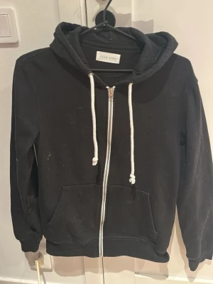 Svart hoodie med dragkedja Pier One - Svart hoodie från Pier One med dragkedja framtill, vita dragsnören och två fickor. Klassisk modell med huva och ribbade muddar vid ärmslut och nederkant. Perfekt för en avslappnad och stilren look.