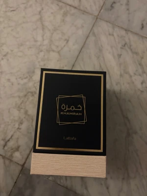 Lattafa Khamrah Eau de Parfum - Lattafa Khamrah Eau de Parfum i en lyxig svart och guld box. Flaskan är fyrkantig med ett elegant mönstrat lock i genomskinligt glas och bärnstensfärgad vätska. Designen känns exklusiv och modern, perfekt för dig som gillar unika dofter och snygga flaskor.