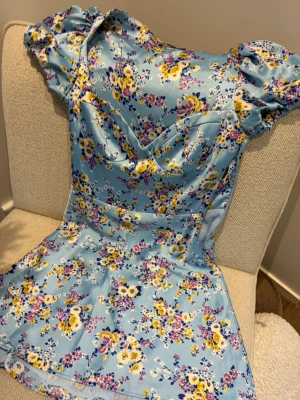 Blommig blå kortklänning med puffärm - Supersöt kort klänning i ljusblått med blommigt mönster i gult, vitt och lila. Klänningen har puffärmar och en v-ringad omlottliknande överdel. Materialet är glansigt och känns som satin eller polyester. Perfekt för sommarens alla tillfällen!