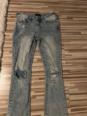 Ljusblå bootcut jeans från True Religion - Säljer ett par ljusblå bootcut jeans från True Religion med slitningar och hål på knäna. Jeansen har klassiska bakfickor med lock och silverfärgade knappar samt en tvättad look. Har vinröda detaljer som gör det lätt att matcha till en vinröd tröja exempelvis.  Perfekta för en avslappnad och trendig stil.
