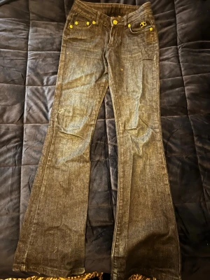 Mörkblåa jeans - Säljer ett par mörkblåa jeans, med utsvängda ben och snygga guldiga detaljer. Bakfickorna har stora broderade loggor i guld och nitarna är också guldiga . Klassisk femficksmodell i denim med låg midja.