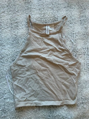 Beige crop top från H&M Divided - Snygg beige crop top från H&M Divided hängande med smala ax bygelband och hög halsringning. Tillverkad i mjuk bomullsmix som känns skön mot huden. Perfekt till jeans eller kjol för en enkel och clean look.