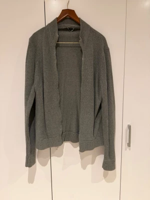 Zara Grå stickad kofta  - En stilren grå stickad kofta med dragkedja framtill och ribbstickade muddar. Koftan har en rak passform och hög krage, perfekt för lager-på-lager. Materialet är mjukt och ger en avslappnad vibe.