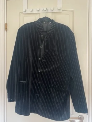 Svart sammetskavaj med vita ränder från Mario Conti - Snygg svart sammetskavaj med tunna vita kritstrecksränder och ståkrage. Kavajen har tre knappar framtill, två fickor och en bröstficka. Perfekt för dig som vill sticka ut med en unik och trendig look.