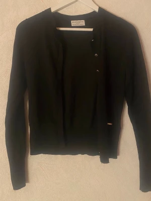 Svart kofta från Holly & White - Stilren svart kofta från Holly & White med rund halsringning och knäppning framtill. Långärmad modell i mjukt, tunt material som passar perfekt till lager på lager. Liten metallbricka med logga vid nederkanten ger en snygg detalj.