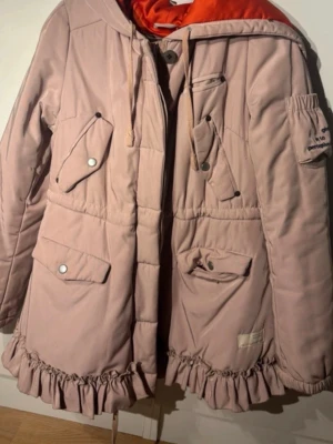 Rosa parkas från Odd Molly - Supersnygg rosa parkas från Odd Molly med volangkant nedtill och orange foder. Unik och väldigt populär under stora Odd Molly eran. Jackan har flera fickor med lock och tryckknappar, dragsko i midjan och huva. 
