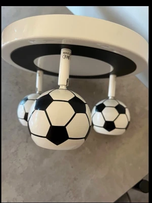 Taklampa med fotbollstema - Säljer en unik taklampa med tre lampor formade som klassiska svartvita fotbollar. Lampan är rund och vit med svarta detaljer och passar perfekt för dig som gillar sportig inredning. Tillverkad i metall och plast. Perfekt till barnrummet eller spelrummet!