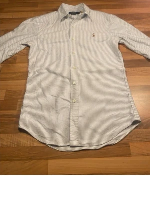 Randig skjorta från Ralph Lauren - Klassisk skjorta från Ralph Lauren i vitt med tunna blå ränder. Skjortan har lång ärm, knappar framtill och en liten broderad logga på bröstet. Tillverkad i bomull och har en tidlös, normal passform.