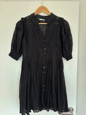 Svart spetsklänning från Mango - Svart kortklänning från Mango med fina spetsdetaljer och knappar framtill. Klänningen har trekvartsärm, hög hals med spets och ett lätt, luftigt tyg i bomull. Perfekt för dig som gillar romantiska detaljer och en klassisk look.