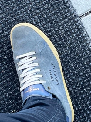 Blå Saint Laurent sneakers i mocka - Snygga blå sneakers från Saint Laurent i mjuk mocka med vit sula och vita skosnören. Märket syns tydligt på sidan och på plösen. Skorna har en klassisk låg profil och en stilren design som passar till jeans eller streetwear.